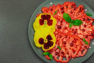 Aile yadigarı Coeur de Boeuf domatesi fesleğenli ve yenebilir çiçekli koyu tabakta. Temiz yemek, vejetaryen yaşam tarzı ve tarladan sofraya yiyecek konsepti.