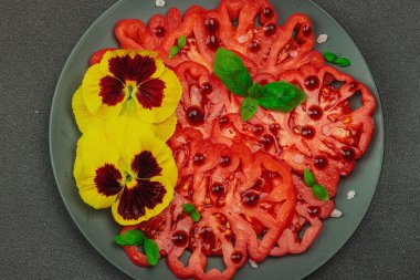 Aile yadigarı Coeur de Boeuf domatesi fesleğenli ve yenebilir çiçekli koyu tabakta. Temiz yemek, vejetaryen yaşam tarzı ve tarladan sofraya yiyecek konsepti.