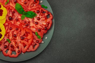 Aile yadigarı Coeur de Boeuf domatesi fesleğenli ve yenebilir çiçekli koyu tabakta. Temiz yemek, vejetaryen yaşam tarzı ve tarladan sofraya yiyecek konsepti.