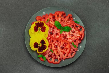 Aile yadigarı Coeur de Boeuf domatesi fesleğenli ve yenebilir çiçekli koyu tabakta. Temiz yemek, vejetaryen yaşam tarzı ve tarladan sofraya yiyecek konsepti.