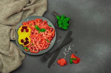 Aile yadigarı Coeur de Boeuf domatesi fesleğenli ve yenebilir çiçekli koyu tabakta. Temiz yemek, vejetaryen yaşam tarzı ve tarladan sofraya yiyecek konsepti.