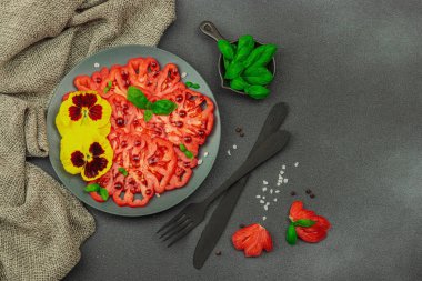 Aile yadigarı Coeur de Boeuf domatesi fesleğenli ve yenebilir çiçekli koyu tabakta. Temiz yemek, vejetaryen yaşam tarzı ve tarladan sofraya yiyecek konsepti.