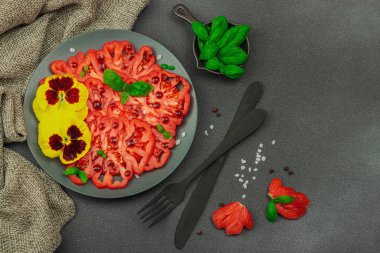 Aile yadigarı Coeur de Boeuf domatesi fesleğenli ve yenebilir çiçekli koyu tabakta. Temiz yemek, vejetaryen yaşam tarzı ve tarladan sofraya yiyecek konsepti.