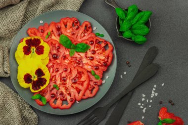 Aile yadigarı Coeur de Boeuf domatesi fesleğenli ve yenebilir çiçekli koyu tabakta. Temiz yemek, vejetaryen yaşam tarzı ve tarladan sofraya yiyecek konsepti.