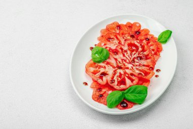 Dilimlenmiş Coeur de Boeuf domatesi, balzamik damlalar ve oval tabakta fesleğen yaprakları. Vejetaryen dostu, aile yadigarı, taş beton arka planda bir çiftlikten bir masaya estetik.