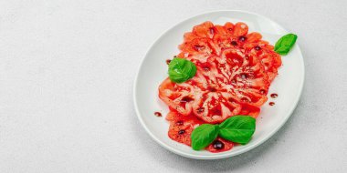 Dilimlenmiş Coeur de Boeuf domatesi, balzamik damlalar ve oval tabakta fesleğen yaprakları. Vejetaryen dostu, aile yadigarı, taş beton arka planda bir çiftlikten bir masaya estetik.