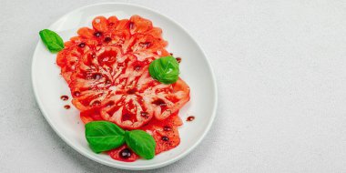 Dilimlenmiş Coeur de Boeuf domatesi, balzamik damlalar ve oval tabakta fesleğen yaprakları. Vejetaryen dostu, aile yadigarı, taş beton arka planda bir çiftlikten bir masaya estetik.