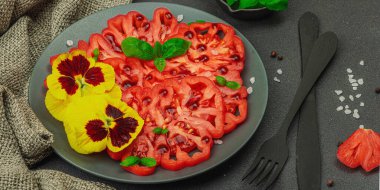 Aile yadigarı Coeur de Boeuf domatesi fesleğenli ve yenebilir çiçekli koyu tabakta. Temiz yemek, vejetaryen yaşam tarzı ve kırsal sunum, afiş formatlı çiftlikten sofraya yiyecek konsepti