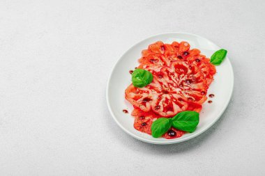 Dilimlenmiş Coeur de Boeuf domatesi, balzamik damlalar ve oval tabakta fesleğen yaprakları. Vejetaryen dostu, aile yadigarı, taş beton arka planda bir çiftlikten bir masaya estetik.