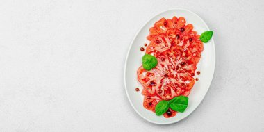 Dilimlenmiş Coeur de Boeuf domatesi, balzamik damlalar ve oval tabakta fesleğen yaprakları. Vejetaryen dostu, aile yadigarı, taş beton arka planda bir çiftlikten bir masaya estetik.