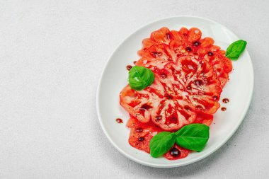 Dilimlenmiş Coeur de Boeuf domatesi, balzamik damlalar ve oval tabakta fesleğen yaprakları. Vejetaryen dostu, aile yadigarı, taş beton arka planda bir çiftlikten bir masaya estetik.