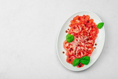 Dilimlenmiş Coeur de Boeuf domatesi, balzamik damlalar ve oval tabakta fesleğen yaprakları. Vejetaryen dostu, aile yadigarı, taş beton arka planda bir çiftlikten bir masaya estetik.