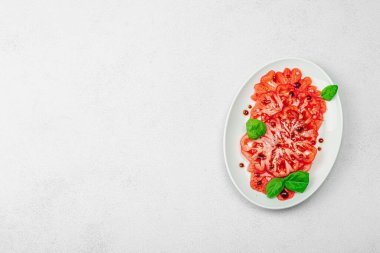 Dilimlenmiş Coeur de Boeuf domatesi, balzamik damlalar ve oval tabakta fesleğen yaprakları. Vejetaryen dostu, aile yadigarı, taş beton arka planda bir çiftlikten bir masaya estetik.