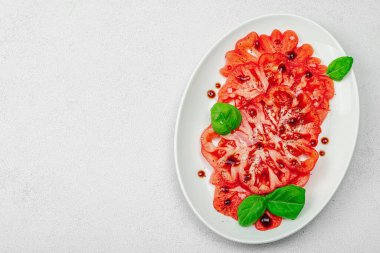 Dilimlenmiş Coeur de Boeuf domatesi, balzamik damlalar ve oval tabakta fesleğen yaprakları. Vejetaryen dostu, aile yadigarı, taş beton arka planda bir çiftlikten bir masaya estetik.