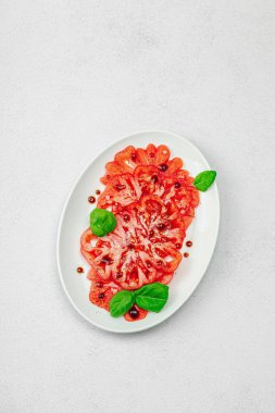 Dilimlenmiş Coeur de Boeuf domatesi, balzamik damlalar ve oval tabakta fesleğen yaprakları. Vejetaryen dostu, aile yadigarı, taş beton arka planda bir çiftlikten bir masaya estetik.