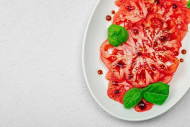 Dilimlenmiş Coeur de Boeuf domatesi, balzamik damlalar ve oval tabakta fesleğen yaprakları. Vejetaryen dostu, aile yadigarı, taş beton arka planda bir çiftlikten bir masaya estetik.