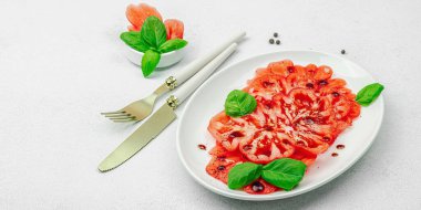 Dilimlenmiş Coeur de Boeuf domatesi, balzamik damlalar ve oval tabakta fesleğen yaprakları. Vejetaryen dostu, aile yadigarı, taş beton arka planda bir çiftlikten bir masaya estetik.