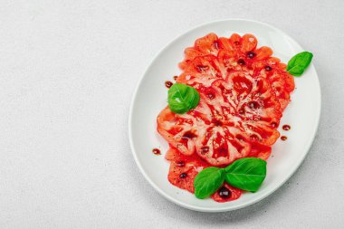 Dilimlenmiş Coeur de Boeuf domatesi, balzamik damlalar ve oval tabakta fesleğen yaprakları. Vejetaryen dostu, aile yadigarı, taş beton arka planda bir çiftlikten bir masaya estetik.