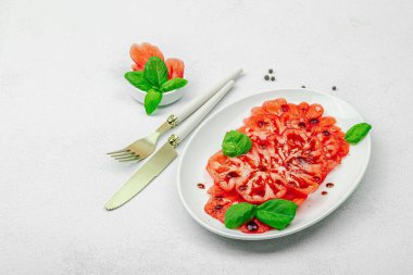 Dilimlenmiş Coeur de Boeuf domatesi, balzamik damlalar ve oval tabakta fesleğen yaprakları. Vejetaryen dostu, aile yadigarı, taş beton arka planda bir çiftlikten bir masaya estetik.