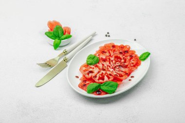 Dilimlenmiş Coeur de Boeuf domatesi, balzamik damlalar ve oval tabakta fesleğen yaprakları. Vejetaryen dostu, aile yadigarı, taş beton arka planda bir çiftlikten bir masaya estetik.