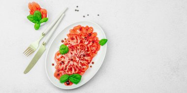 Dilimlenmiş Coeur de Boeuf domatesi, balzamik damlalar ve oval tabakta fesleğen yaprakları. Vejetaryen dostu, aile yadigarı, taş beton arka planda bir çiftlikten bir masaya estetik.