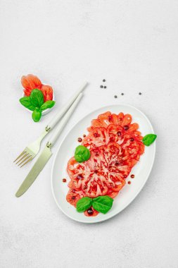 Dilimlenmiş Coeur de Boeuf domatesi, balzamik damlalar ve oval tabakta fesleğen yaprakları. Vejetaryen dostu, aile yadigarı, taş beton arka planda bir çiftlikten bir masaya estetik.