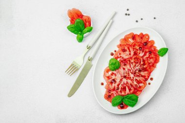 Dilimlenmiş Coeur de Boeuf domatesi, balzamik damlalar ve oval tabakta fesleğen yaprakları. Vejetaryen dostu, aile yadigarı, taş beton arka planda bir çiftlikten bir masaya estetik.