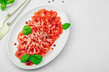 Dilimlenmiş Coeur de Boeuf domatesi, balzamik damlalar ve oval tabakta fesleğen yaprakları. Vejetaryen dostu, aile yadigarı, taş beton arka planda bir çiftlikten bir masaya estetik.