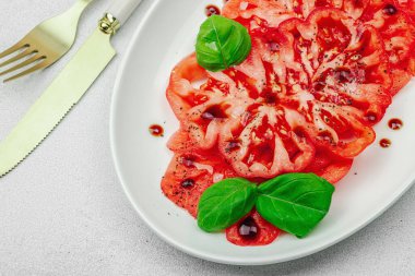 Dilimlenmiş Coeur de Boeuf domatesi, balzamik damlalar ve oval tabakta fesleğen yaprakları. Vejetaryen dostu, aile yadigarı, taş beton arka planda bir çiftlikten bir masaya estetik.