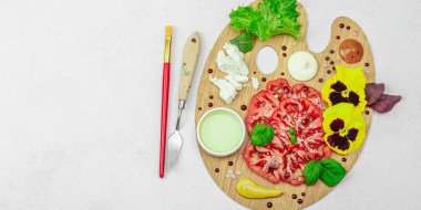 Dilimlenmiş aile yadigarı domates, feta, otlar, yenilebilir çiçekler, ve ahşap palette soslar, fırça ve palet bıçağıyla şekillendirilmiş. Hafif taş arkaplan, pankart biçimi