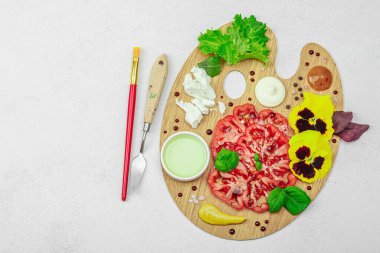 Dilimlenmiş aile yadigarı domates, feta, otlar, yenilebilir çiçekler, ve ahşap palette soslar, fırça ve palet bıçağıyla şekillendirilmiş. Hafif taş arkaplan, üst görünüm