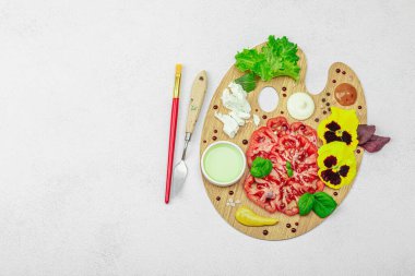 Dilimlenmiş aile yadigarı domates, feta, otlar, yenilebilir çiçekler, ve ahşap palette soslar, fırça ve palet bıçağıyla şekillendirilmiş. Hafif taş arkaplan, üst görünüm