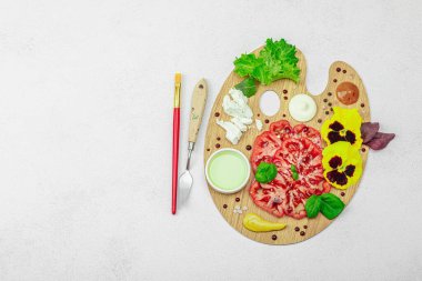Dilimlenmiş aile yadigarı domates, feta, otlar, yenilebilir çiçekler, ve ahşap palette soslar, fırça ve palet bıçağıyla şekillendirilmiş. Hafif taş arkaplan, üst görünüm
