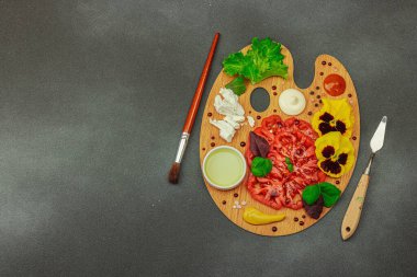 Dilimlenmiş aile yadigarı domates, feta, otlar, yenilebilir çiçekler, ve ahşap palette soslar, fırça ve palet bıçağıyla şekillendirilmiş. Koyu taş zemin, üst görünüm