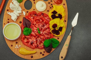 Dilimlenmiş aile yadigarı domates, feta, otlar, yenilebilir çiçekler, ve ahşap palette soslar, fırça ve palet bıçağıyla şekillendirilmiş. Koyu taş zemin, üst görünüm