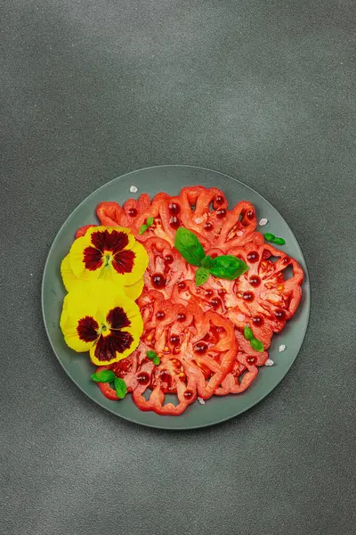 Aile yadigarı Coeur de Boeuf domatesi fesleğenli ve yenebilir çiçekli koyu tabakta. Temiz yemek, vejetaryen yaşam tarzı ve tarladan sofraya yiyecek konsepti.