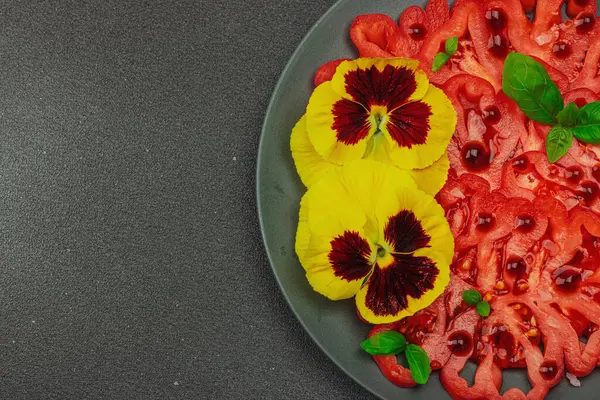 Aile yadigarı Coeur de Boeuf domatesi fesleğenli ve yenebilir çiçekli koyu tabakta. Temiz yemek, vejetaryen yaşam tarzı ve tarladan sofraya yiyecek konsepti.