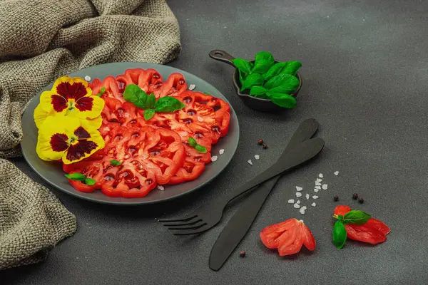 Aile yadigarı Coeur de Boeuf domatesi fesleğenli ve yenebilir çiçekli koyu tabakta. Temiz yemek, vejetaryen yaşam tarzı ve kırsal sunumlu tarladan sofraya yiyecek konsepti, kopyalama alanı