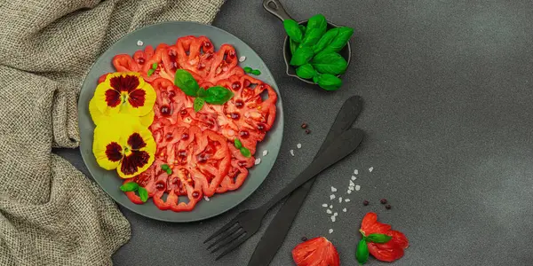 Aile yadigarı Coeur de Boeuf domatesi fesleğenli ve yenebilir çiçekli koyu tabakta. Temiz yemek, vejetaryen yaşam tarzı ve kırsal sunum, afiş formatlı çiftlikten sofraya yiyecek konsepti