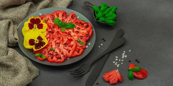 Aile yadigarı Coeur de Boeuf domatesi fesleğenli ve yenebilir çiçekli koyu tabakta. Temiz yemek, vejetaryen yaşam tarzı ve kırsal sunum, afiş formatlı çiftlikten sofraya yiyecek konsepti