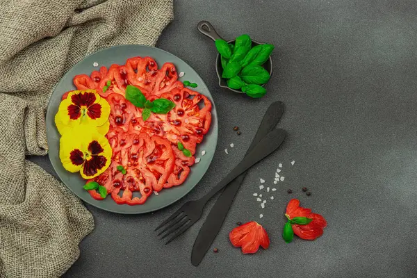 Aile yadigarı Coeur de Boeuf domatesi fesleğenli ve yenebilir çiçekli koyu tabakta. Temiz yemek, vejetaryen yaşam tarzı ve tarladan sofraya yiyecek konsepti.