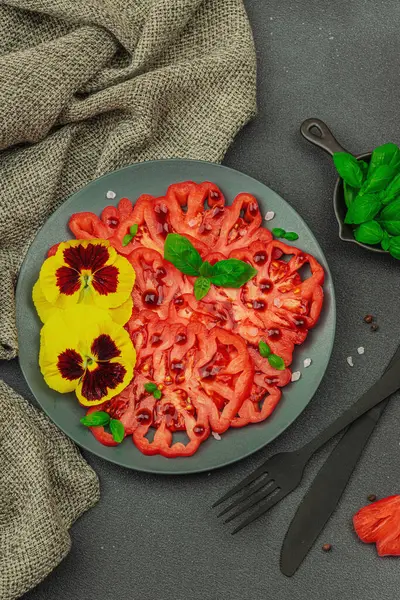 Aile yadigarı Coeur de Boeuf domatesi fesleğenli ve yenebilir çiçekli koyu tabakta. Temiz yemek, vejetaryen yaşam tarzı ve tarladan sofraya yiyecek konsepti.