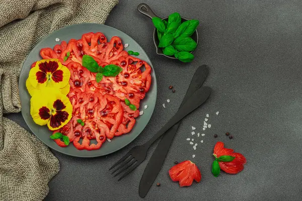 Aile yadigarı Coeur de Boeuf domatesi fesleğenli ve yenebilir çiçekli koyu tabakta. Temiz yemek, vejetaryen yaşam tarzı ve tarladan sofraya yiyecek konsepti.