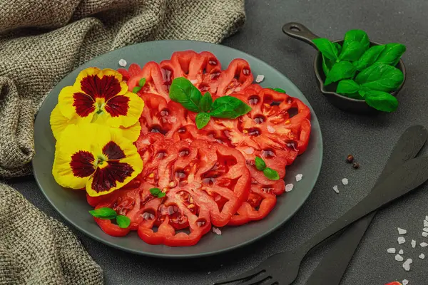 Aile yadigarı Coeur de Boeuf domatesi fesleğenli ve yenebilir çiçekli koyu tabakta. Temiz yemek, vejetaryen yaşam tarzı ve kırsal kesimde sunumlu tarladan sofraya yiyecek konsepti.