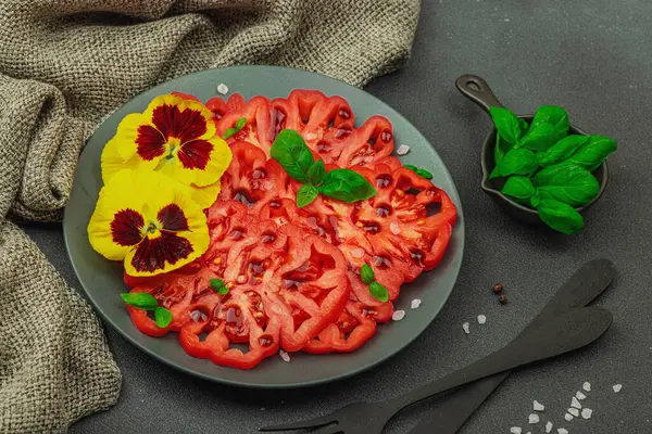 Aile yadigarı Coeur de Boeuf domatesi fesleğenli ve yenebilir çiçekli koyu tabakta. Temiz yemek, vejetaryen yaşam tarzı ve kırsal kesimde sunumlu tarladan sofraya yiyecek konsepti.