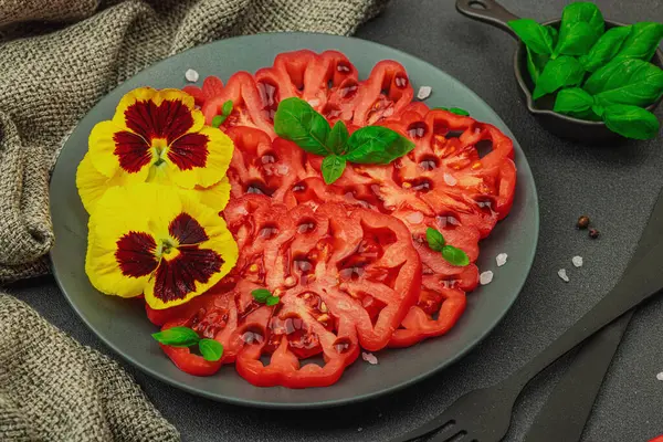 Aile yadigarı Coeur de Boeuf domatesi fesleğenli ve yenebilir çiçekli koyu tabakta. Temiz yemek, vejetaryen yaşam tarzı ve kırsal kesimde sunumlu tarladan sofraya yiyecek konsepti.