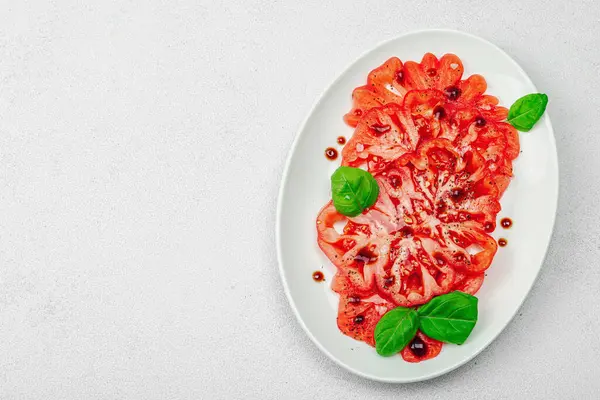 Dilimlenmiş Coeur de Boeuf domatesi, balzamik damlalar ve oval tabakta fesleğen yaprakları. Vejetaryen dostu, aile yadigarı, taş beton arka planda bir çiftlikten bir masaya estetik.