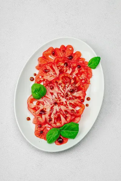 Dilimlenmiş Coeur de Boeuf domatesi, balzamik damlalar ve oval tabakta fesleğen yaprakları. Vejetaryen dostu, aile yadigarı, taş beton arka planda bir çiftlikten bir masaya estetik.