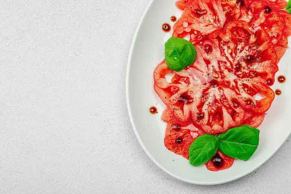 Dilimlenmiş Coeur de Boeuf domatesi, balzamik damlalar ve oval tabakta fesleğen yaprakları. Vejetaryen dostu, aile yadigarı, taş beton arka planda bir çiftlikten bir masaya estetik.