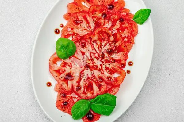 Dilimlenmiş Coeur de Boeuf domatesi, balzamik damlalar ve oval tabakta fesleğen yaprakları. Vejetaryen dostu, aile yadigarı, taş beton arka planda bir çiftlikten bir masaya estetik.